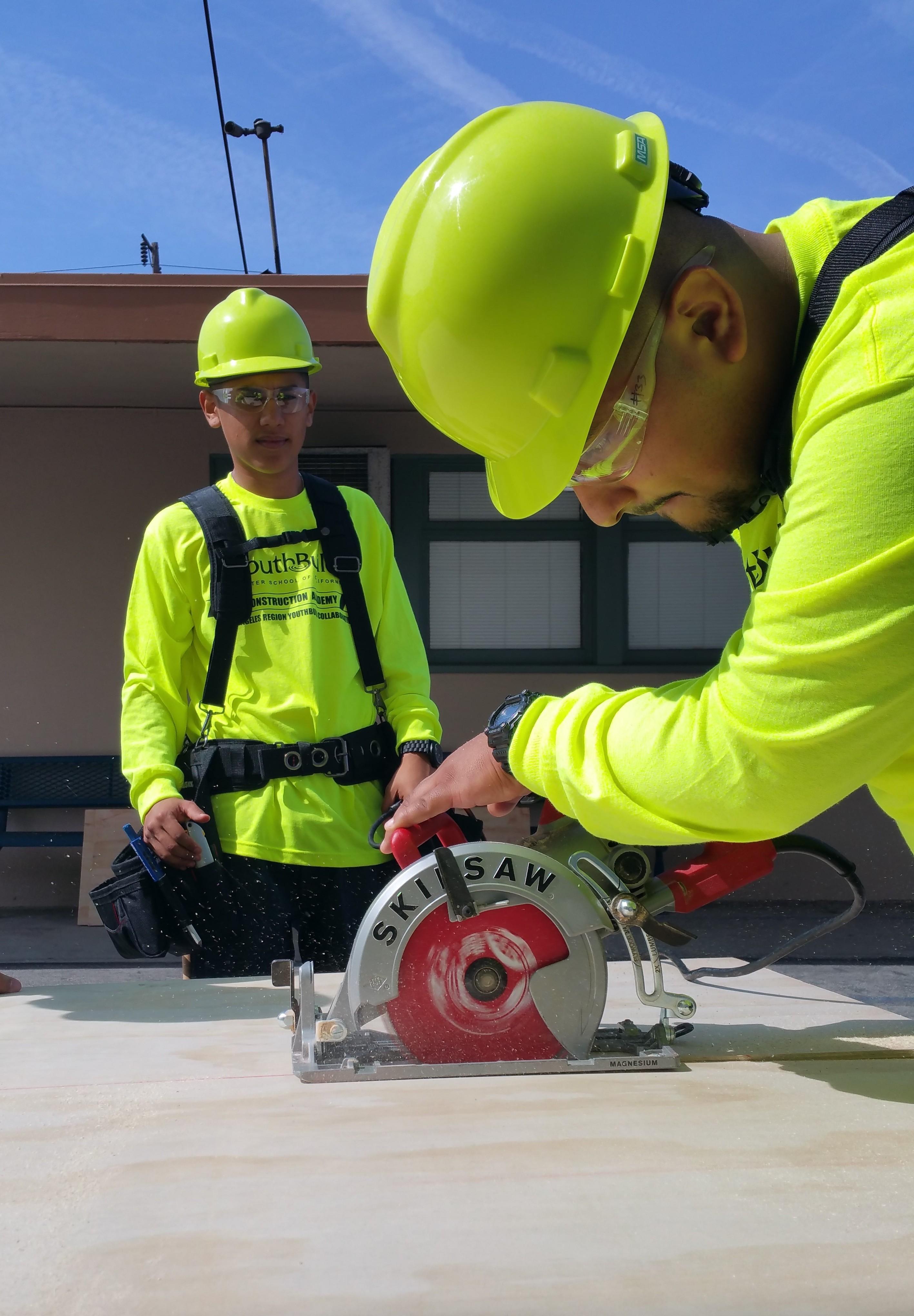 two YouthBuild construction students in hard hats operating a circular saw