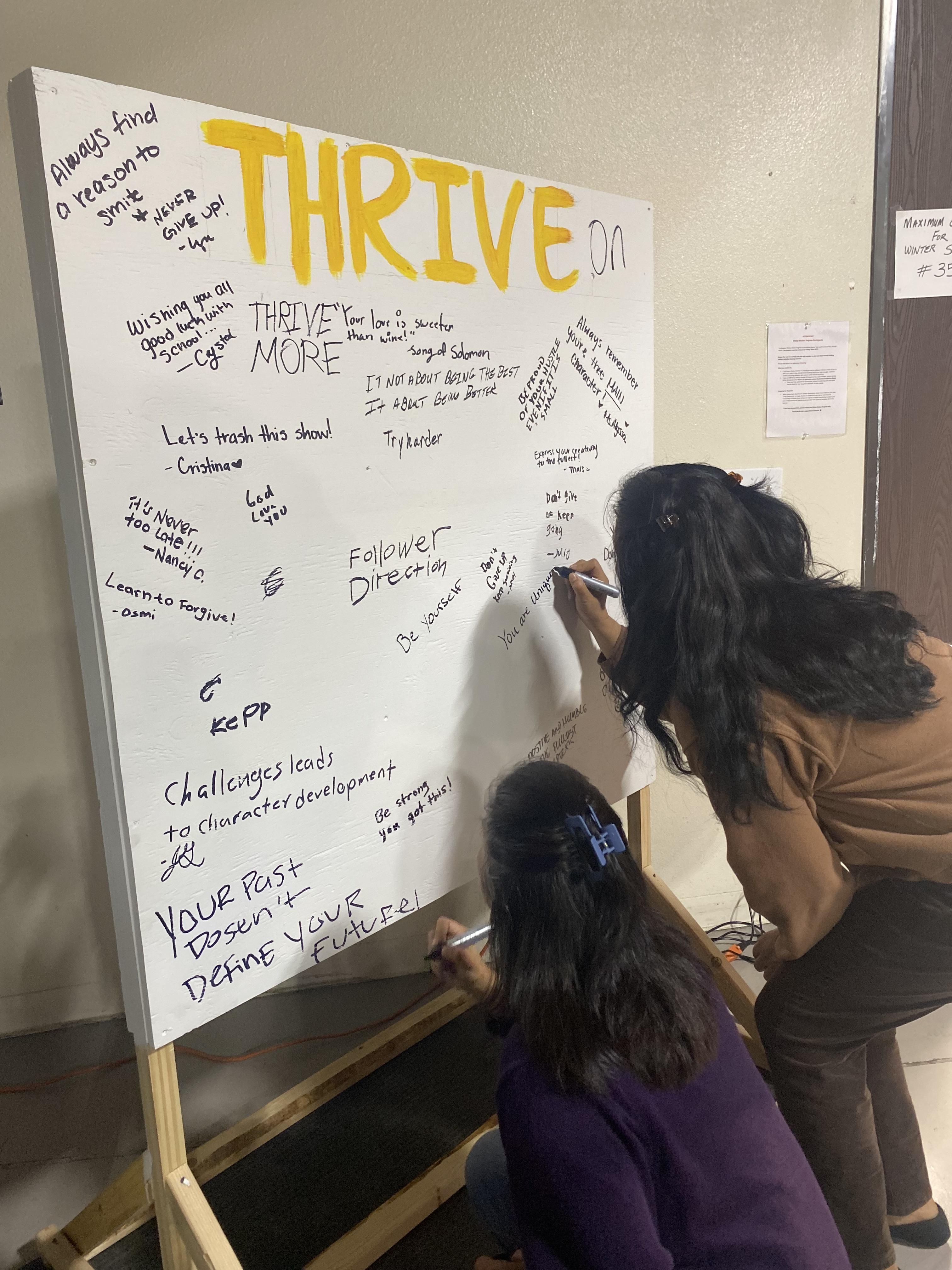 Two women write on the "Thrive" billboard